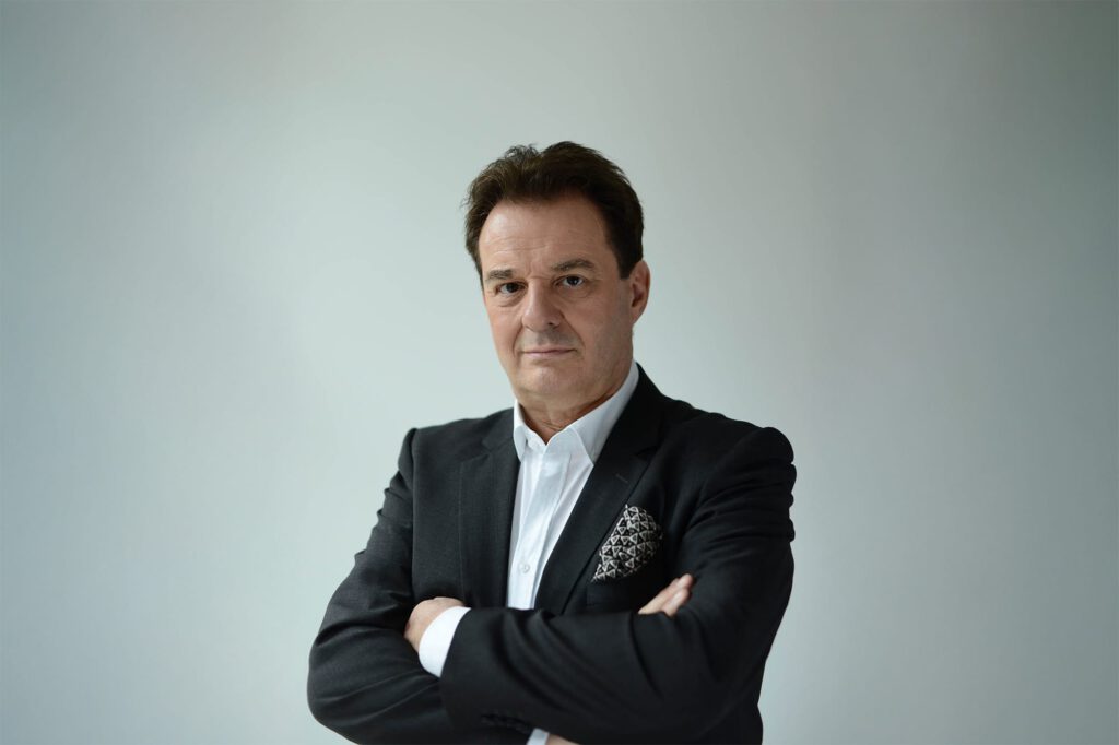 Porträt von Prof. im weißen Kittel; konzentrierter, ruhiger Ausdruck vor neutralem Hintergrund.