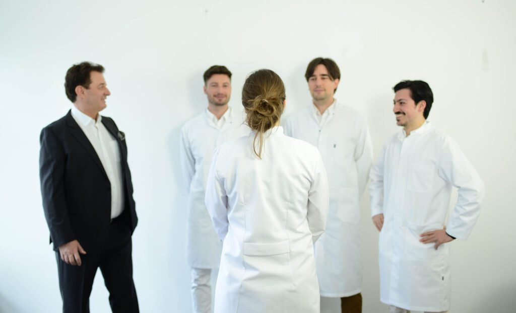 Gruppenfoto von fünf Ärztinnen und Ärzten in weißer Kleidung; das Team steht im Gespräch in heller Praxisumgebung.