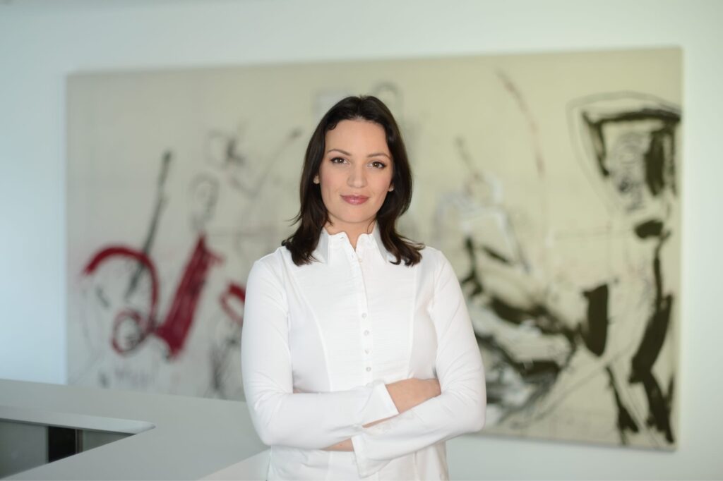 Porträt von K. Fischinger in weißem Kittel, stehend vor Wandkunst in heller Praxis; ruhiger Blick und professionelle Ausstrahlung.